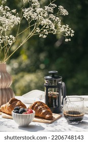 table serving white linen tablecloth, sweet croissants, ripe berry, black coffee in glass french press and flowers in vase in garden on blurred background at summer day