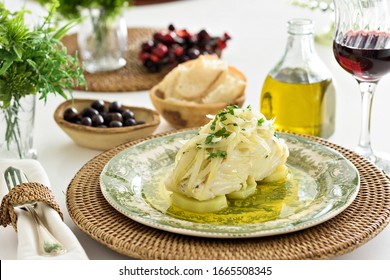 Table set for a dinner, with green decorated portuguese plate, glass of wine and some side dishes. A codfish with potatoes, onion and parsley is being served, an typical portuguese dish.