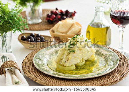 Table set for a dinner, with green decorated portuguese plate, glass of wine and some side dishes. A codfish with potatoes, onion and parsley is being served, an typical portuguese dish.