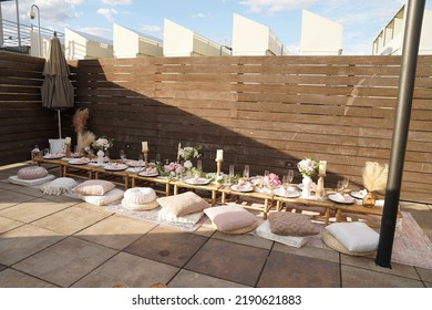 A Table Is Set Up For A Fancy Picnic On Top Of A Residential Rooftop With Pillow Cushions And Nice Plates, Utensils And Stemware