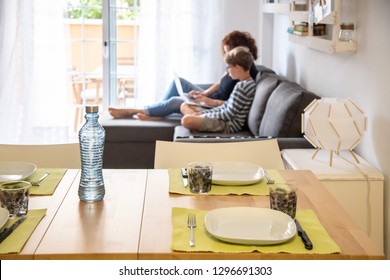 Table set for lunch, white ceramic dishes and decorated glasses with leaves placemats green. Mom and son using a computer sitting on the sofa in the background. Family scene at home mother and son