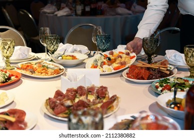 Table Is Set With Variety Of Gourmet Dishes And Appetizers For Banquet. Waiter's Hand Putting Plate On Table. Selective Focus