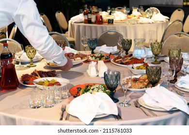 Table Is Set With Variety Of Gourmet Dishes And Appetizers For Banquet. Waiter's Hand Putting Plate On Table. Selective Focus