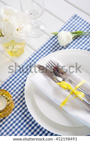 Table setting with blue checkered tablecloth, white napkin, silverware, yellow decoration and flowers