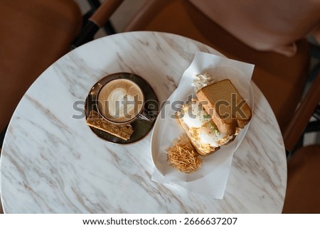 Table setting in a cozy cafe, featuring a single sandwich on toasted bread with a hearty filling and a cup of coffee with froth on a saucer, highlighting a classic breakfast or lunch moment.