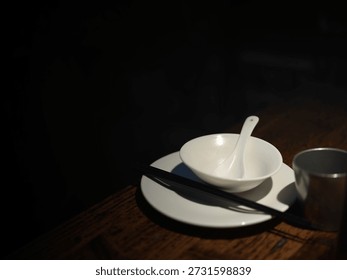 A Table Setting in Dim Lighting Featuring a White Bowl, Spoon, Chopsticks, and Metal Cup on a Wooden Surface