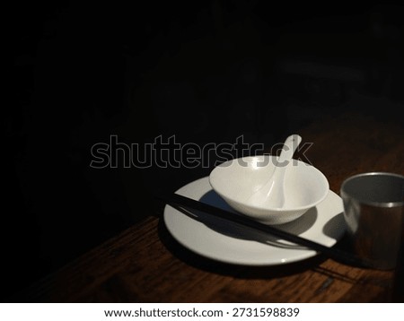 A Table Setting in Dim Lighting Featuring a White Bowl, Spoon, Chopsticks, and Metal Cup on a Wooden Surface