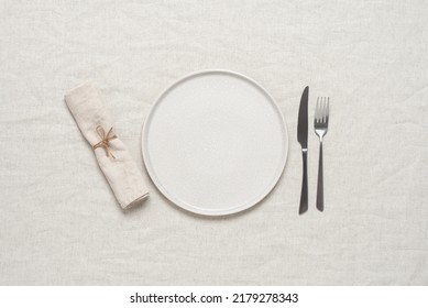 Table setting. Empty beige plate, cutlery and beige linen napkin on beige linen tablecloth. Top view, flat lay. Textured object, selective focus.