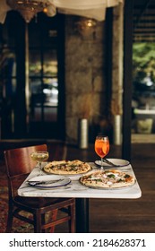 Table Setting In A Pizzeria. Two Italian Pizzas With Burnt Sides And Cold Summer Cocktails On A Marble Square Table In A Cafe Restaurant.