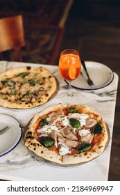 Table Setting In A Pizzeria. Two Italian Pizzas With Burnt Sides And A Cold Summer Cocktail On A Marble Table In A Cafe. Pizza With Sausage, Cheese And Basil.