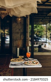 Table Setting In A Pizzeria. Two Italian Pizzas With Burnt Sides And Cold Summer Cocktails On A Marble Square Table In A Cafe Restaurant.