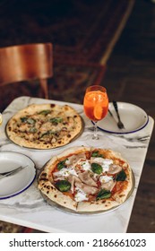 Table Setting In A Pizzeria. Two Italian Pizzas With Burnt Sides And A Cold Summer Cocktail On A Marble Table In A Cafe. Pizza With Sausage, Cheese And Basil.