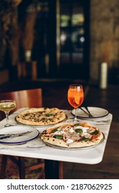 Table Setting In A Pizzeria. Two Italian Pizzas With Burnt Sides And Cold Summer Cocktails On A Marble Square Table In A Cafe Restaurant.