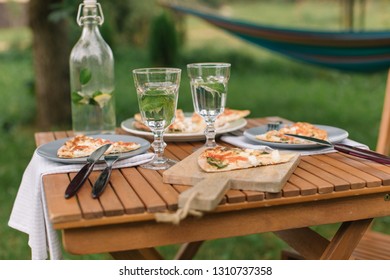Table setting for two at summer terrace. Delicious pizza, glasses of water with lemon and mint, modern crockery on wooden table outdoors. 