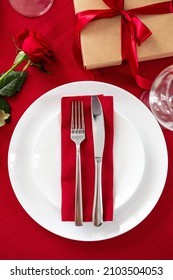 Table Setting For Valentines Day Romantic Dinner. Top View Fork And Knife On Red Napkin On Plate, Rose Flower, Gift Box, Glasses On Red Table.