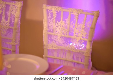 Table setup for a formal dining experience such a wedding or a banquet with focus on a white chair backrest covered in a thin, see-through silky cloth with a purple light delicately reflecting through
