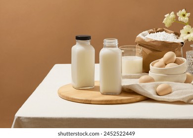 A table with tablecloth features some ingredients for baking such as eggs, flour and milk. A wooden chopping board with two blank label milk bottles displayed on for product promotion - Powered by Shutterstock