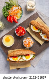 Table Top Flatlay Shot Of Banh Mi - Vietnamese Sandwich With Sausage, Pork, Lettuce, Tomato, Eggs And Arugula On The Light Grey Texture Background. Surrounding By Ingredients