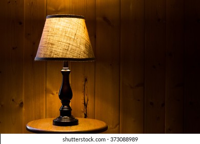 Table Top Lamp With Burlap Shade Lit Over A Wooden Textured Background On A Wooden Table