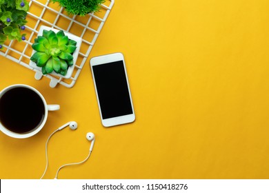 Table Top View Aerial Image Of Accessories Office Desk Background Concept.Flat Lay Of Variety Object The White Mobile Phone & Tree Pot And Coffee With Earphone On Modern Rustic Yellow Paper And Space.