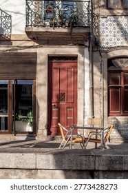 A table and two chairs sit outside a building with a red door, a window with blinds, and a window with blue and white tiles.