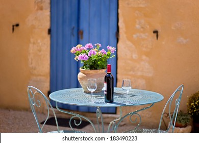 Table For Two Set With Red Wine In Front Of A Rustic Country House. The Wall Is Painted In Ochre  With Wooden Door In Blue.