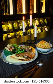 Table With Vietnamese Cooked Meat Food With Lots Of Vegetable And Potato Paste. Three Burning Candles And Wine Rack Int The Background