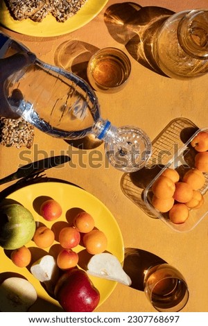 Tablescape on orange background in sharp daylight with fruits in yellow plate: apricots, pear, apple and glasses with alcoholic drinks: cognac and water and hand pouring water from plastic bottle