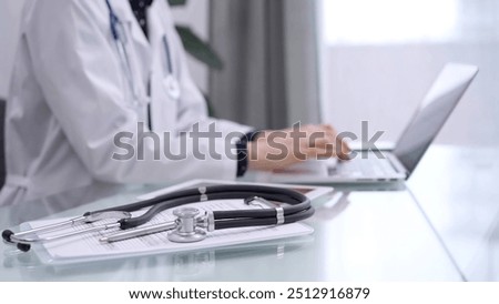 Tablet computer and black stethoscope are on the glass table besides a doctor woman using laptop computer on the glass table. Medicine concept
