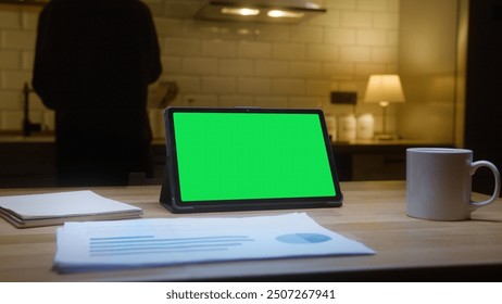 Tablet computer with mock up green screen chromakey display standing on the kitchen table. Anonymous person is preparing something in the background in kitchen at night.