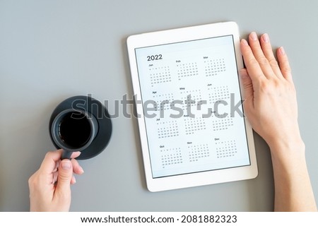 tablet computer with open app of calendar for 2022 year in a womans hands and cup of tea or coffee on a gray background. concept business or to do list goals with technology using. top view, flat lay
