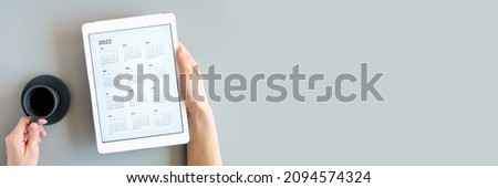 tablet computer with open app of calendar for 2022 year in a woman's hands and cup of tea or coffee on gray background. concept business or to do list goals. top view, flat lay. banner. space for text