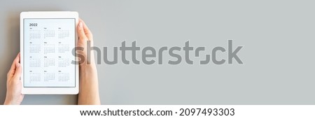 tablet computer with an open app of calendar for 2022 year in womans hands on a gray background. concept business or to do list goals with technology using. top view, flat lay. banner. space for text