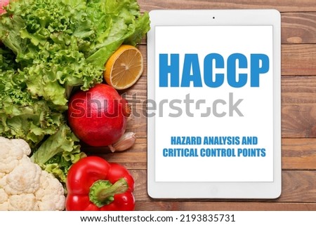 Tablet computer with text HAZARD ANALYSIS AND CRITICAL POINTS and fresh vegetables on wooden background
