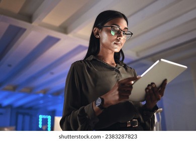 Tablet, designer and serious woman research in business startup office at night on deadline. Technology, creative professional or Indian entrepreneur reading information, email or app in neon company