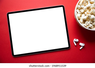 Tablet with empty white screen with wireless earphones and bowl of popcorn next to it. Mockup for video or movie screenshot.