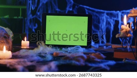 A tablet with a green screen surrounded by candles and cobwebs in a Halloween-themed setting.