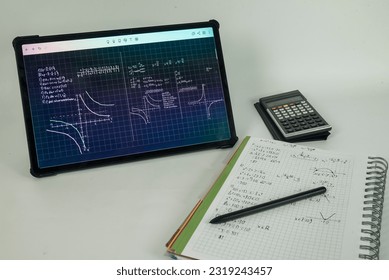 Tablet with math problems displayed. A notebook with notes, a calculator and a pen.
