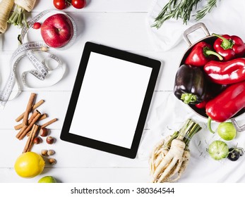 Tablet with a measuring tape, fresh rosemary, lime, lemon, nuts, parsley root, tomatoes, cinnamon, peppers and red apples on a white wooden background top view horizontal - Powered by Shutterstock