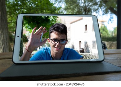 tablet on a wooden table in a garden making a video call with a young man waving at the camera - Shutterstock ID 2043924476