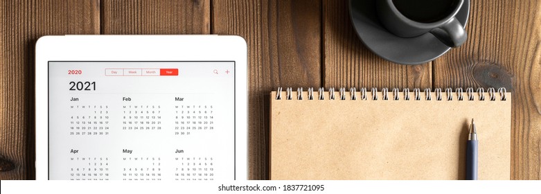 A Tablet With An Open Calendar For 2021 Year, A Cup Of Coffee And A Craft Paper Notebook On A Wooden Boards Table Background. Banner