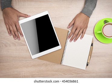 Tablet PC in the hands, a cup of tea and notebook on the desktop. Top view.