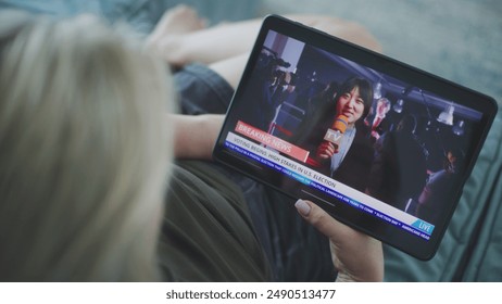 Tablet screen shows playback of breaking news with female Asian correspondent. Caucasian woman watches news broadcast report about US election using tablet computer while lying on sofa. Close Up.