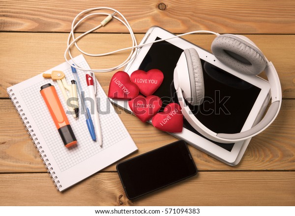 Tablet smartphone touch\
headphones Notepad with pen and marker on wooden background with\
red hearts.