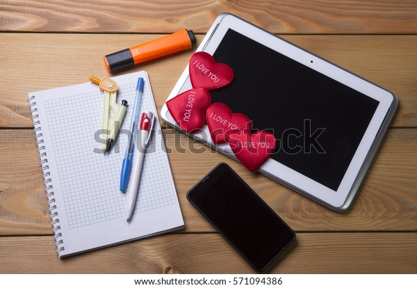 Tablet smartphone touch  Notepad with\
pen and marker on wooden background with red\
hearts.