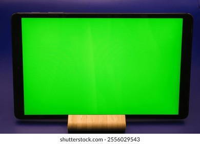 A tablet stands upright on a wooden base displaying a bright green screen. The blue backdrop enhances the vibrant color, making it ideal for video editing, presentations, and digital content creation. - Powered by Shutterstock