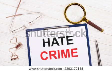A tablet with the text HATE CRIME on white paper, a magnifier, glasses, golden paper clips and a pen lie on a white office table. Top view, flat lay.