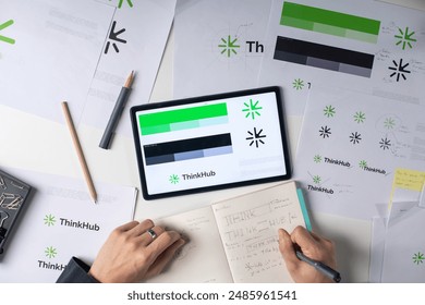 tabletop view of a logo design process, featuring sketches, color swatches, and digital drafts on a tablet. various design tools and materials, including pencils and markers.