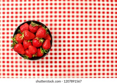 Tabletop view, small black ceramic bowl of strawberries on red chequered gingham tablecloth