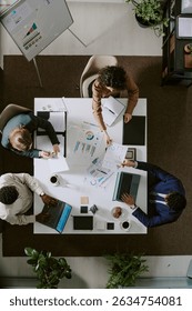 Tabletop view of three professionals working on statistical charts on desk surrounded by plants and equipment. Business meeting involving laptop and documents